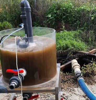 A clear-walled tank of dark brown recycled water on a treatment rig, with hoses and a valve at its base